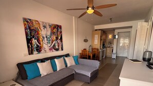 Una sala de estar con un sofá modular, un ventilador de techo y una zona de cocina al fondo.