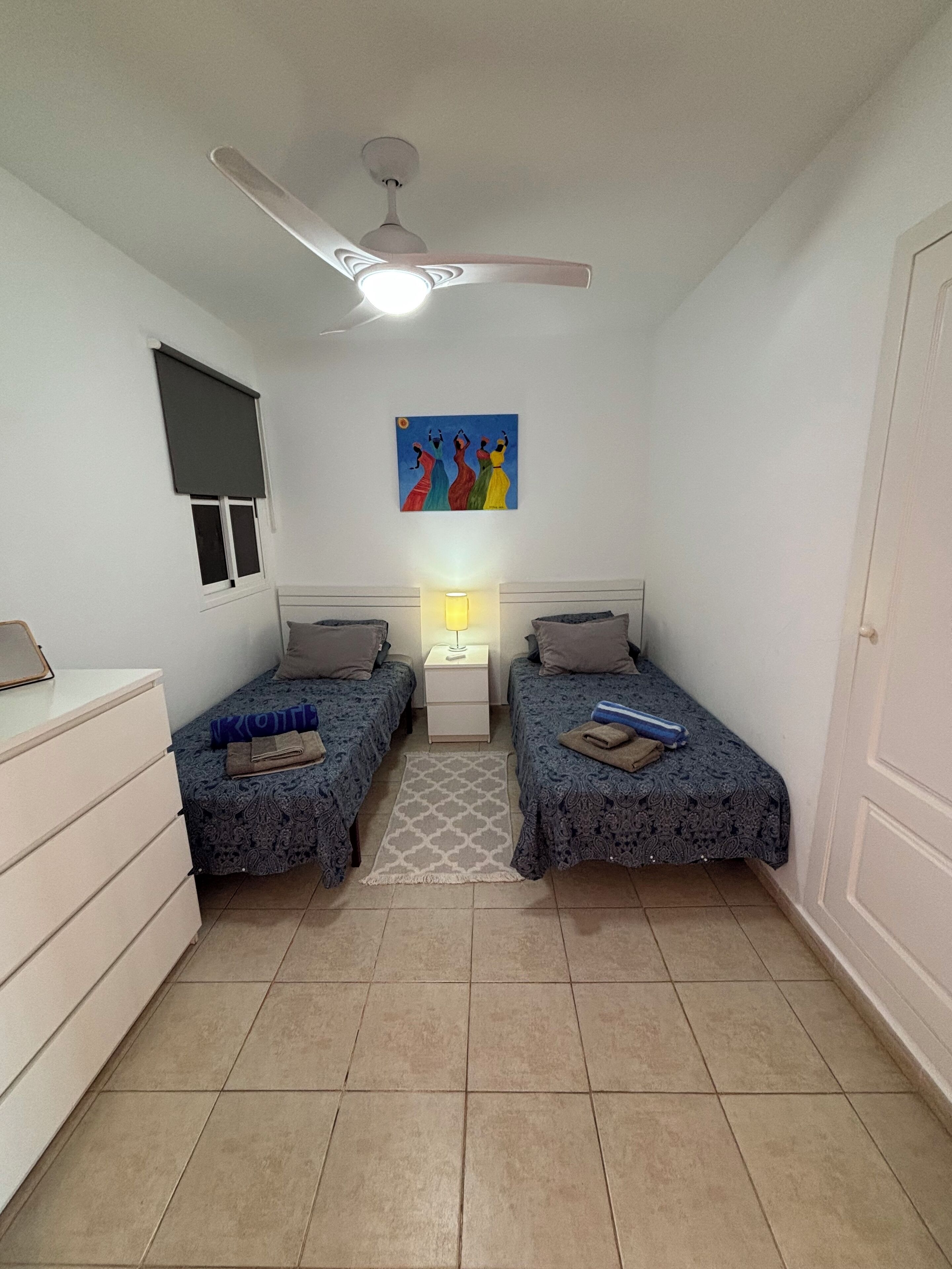 Un dormitorio con dos camas, un ventilador de techo, una mesita de noche con lámpara, una ventana con persianas y un cuadro en la pared.