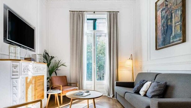 A modern living room with a grey sofa, a round coffee table, a TV, and a large window with curtains. - Luxury Apartment on Valencia Street (Barcelona)
