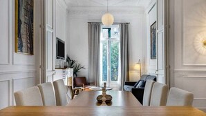 A dining room with a long wooden table, chairs, a chandelier, and a view through a window with curtains. - Luxury Apartment on Valencia Street (Barcelona)