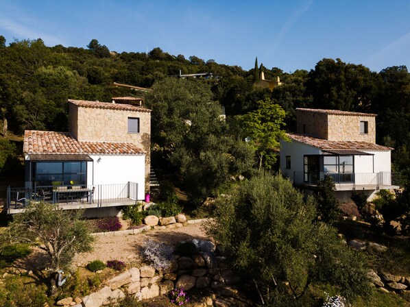 Two stone buildings with tiled roofs, surrounded by dense greenery and hills.