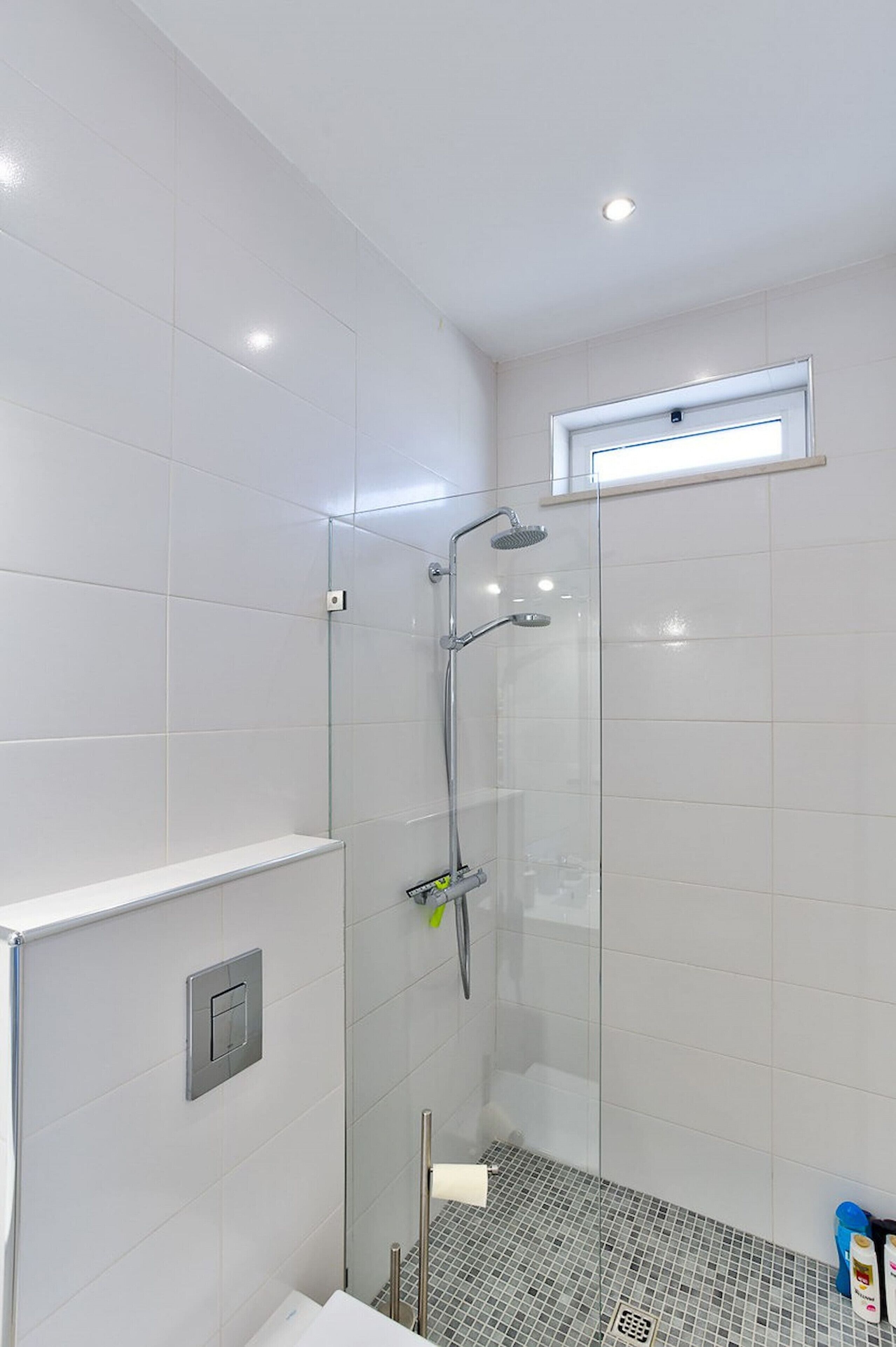 Un bagno moderno con cabina doccia in vetro, pareti rivestite di piastrelle bianche e una piccola finestra sopra la doccia.