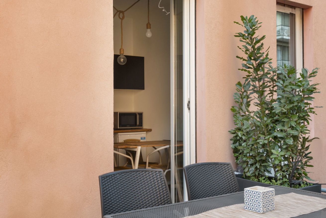 A small outdoor dining area with a table and chairs, a potted plant, and a sliding glass door leading to an interior space with a microwave and a television.