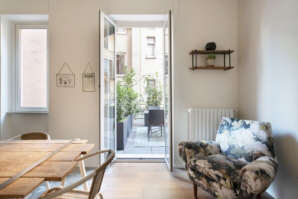 A modern living room with a wooden dining table, a patterned sofa, and a view of a garden through a glass door.