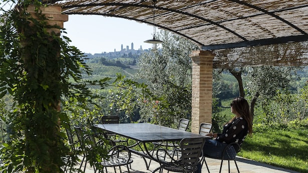 Um pátio externo com mesa e cadeiras, rodeado por vegetação e vista para a cidade.