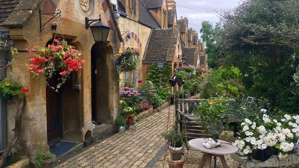 Una estrecha calle adoquinada flanqueada por edificios históricos, cada uno decorado con cestas de flores colgantes y plantas en macetas.