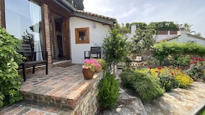 A house with a brick exterior, a wooden door, and a small porch with two chairs. There are potted plants and a variety of flowers in the garden. - Garden House, Ephesus Holiday Rentals (Selçuk)