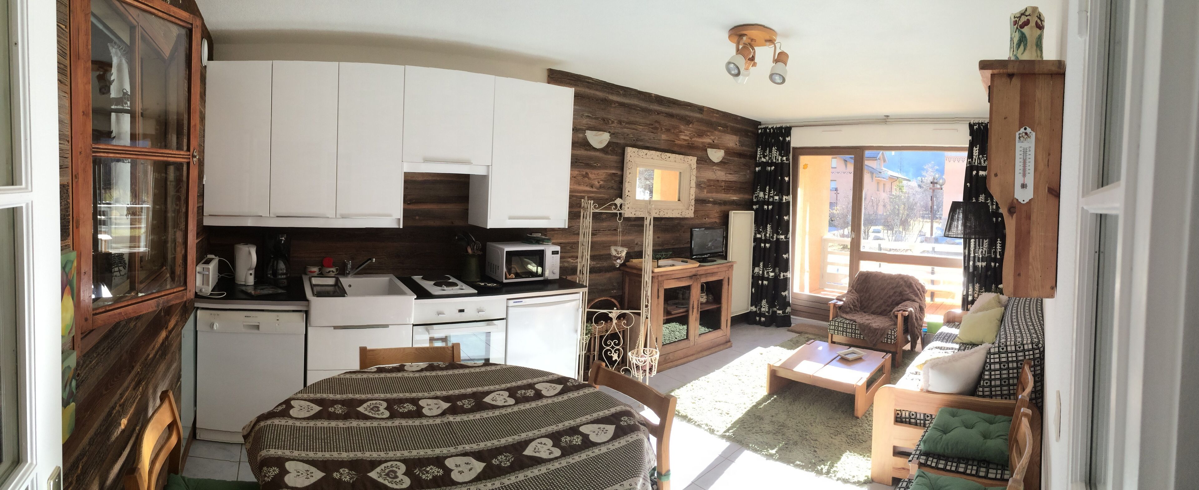 Una cucina compatta con mobili bianchi, un forno a microonde e un fornello. C'è un'area pranzo con tavolo e sedie, e un divano. La stanza presenta una parete d'accento in legno e una finestra con vista esterna.