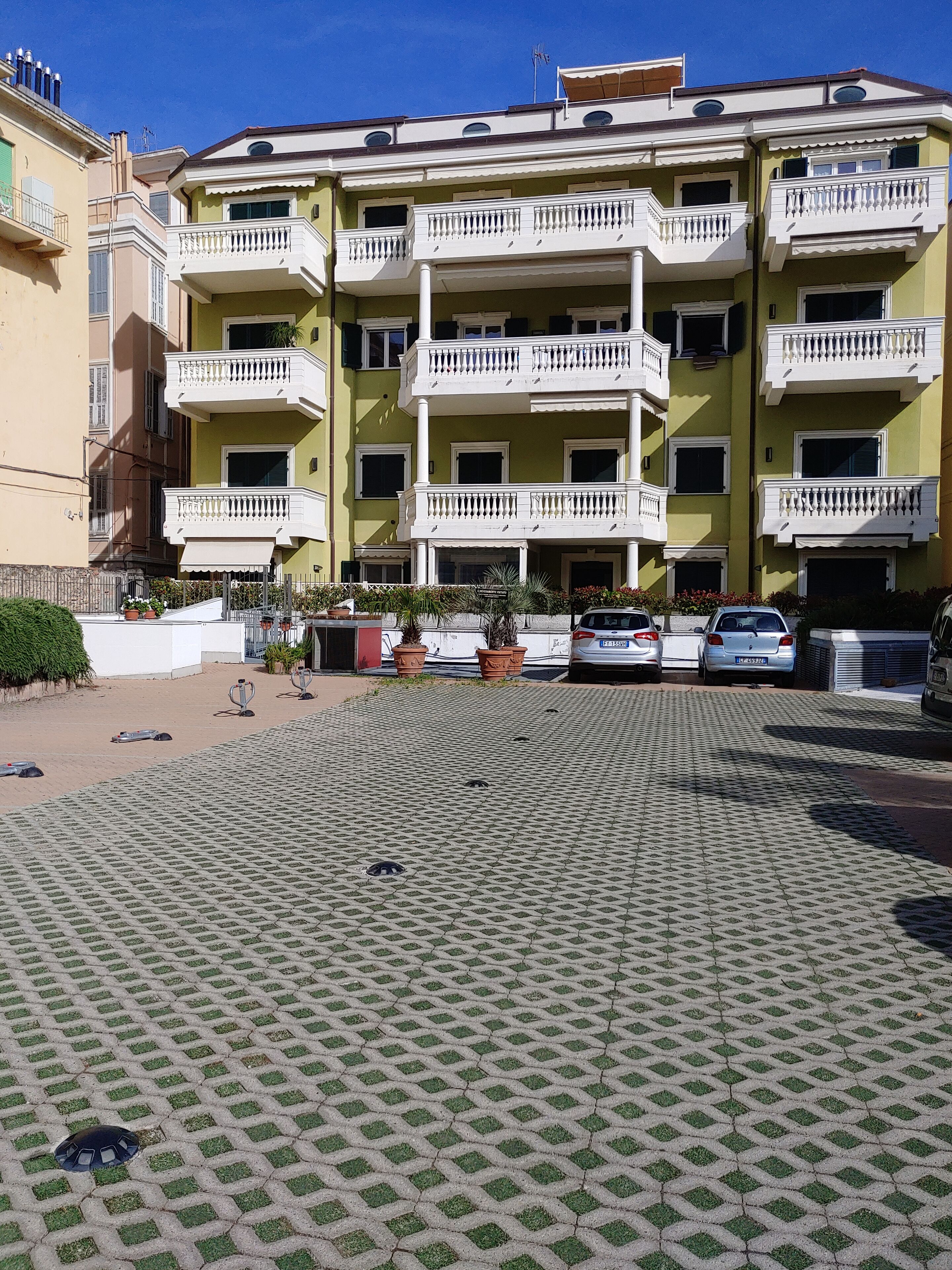 A multi-story residential building with balconies, a paved courtyard, and parked cars.