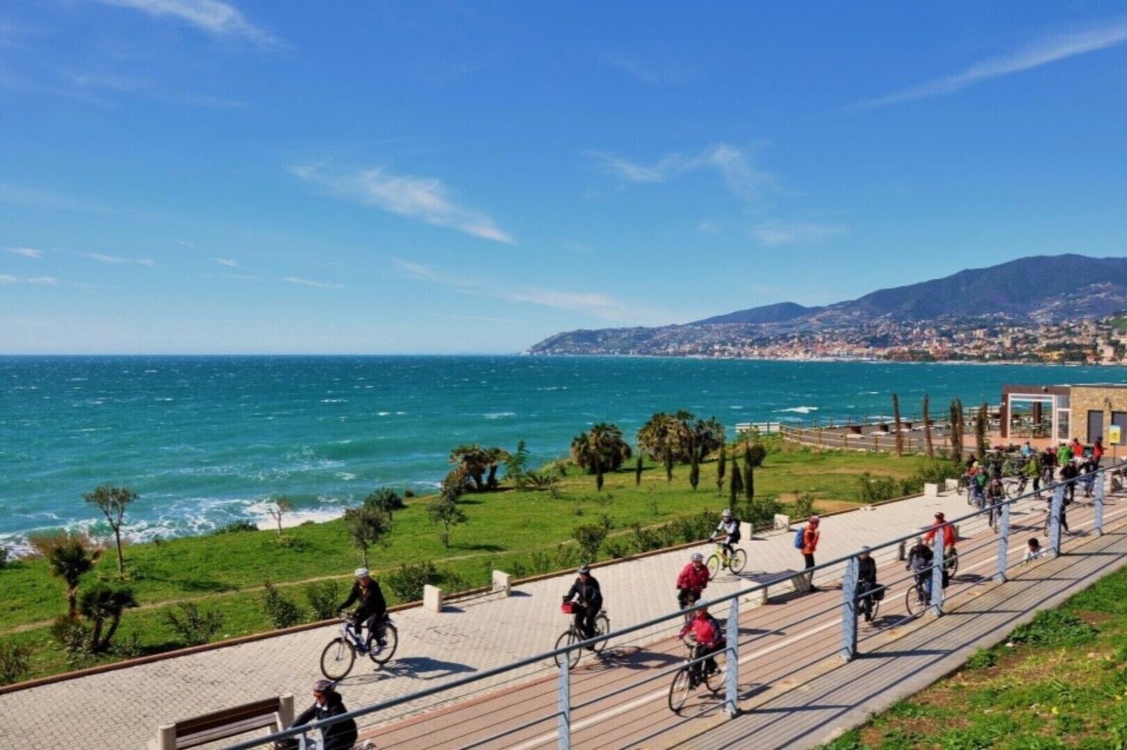 A coastal promenade with cyclists, a clear view of the sea, and a distant cityscape.