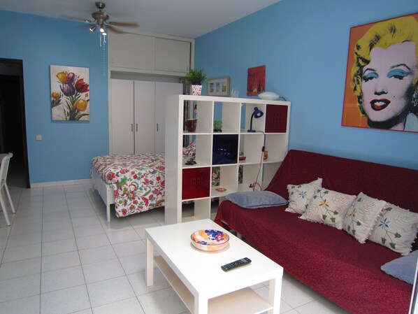 A living room with a red sofa, white coffee table, and a bed with floral bedding. There is a white shelving unit with various items and a ceiling fan.