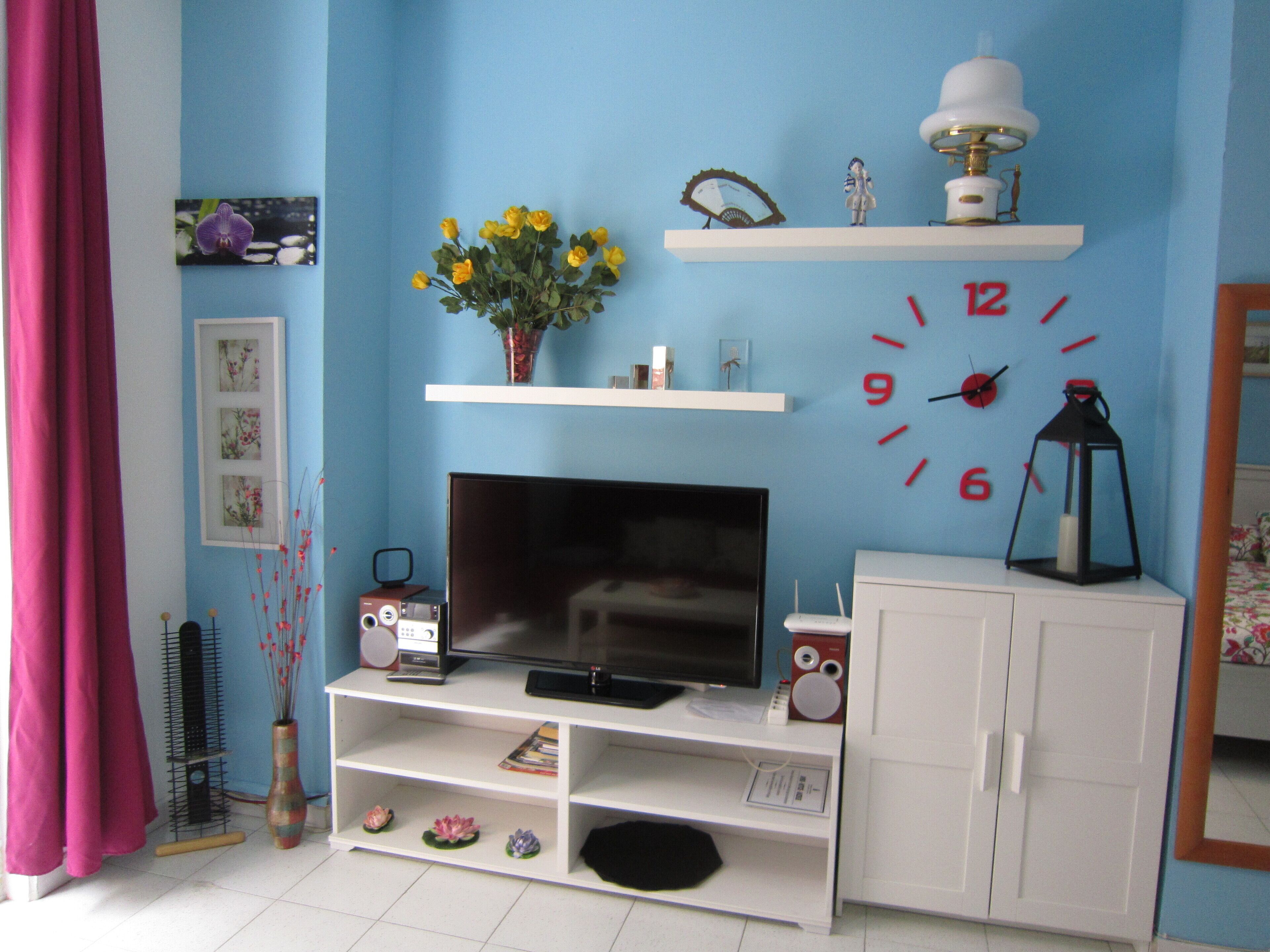 A room with a TV on a white stand, a white cabinet, a clock, and a vase with yellow flowers.