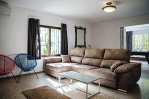 Uma sala de estar com um sofá seccional marrom, uma mesa de centro de vidro, duas cadeiras coloridas, um espelho grande e uma janela com cortinas.