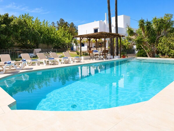 A clear swimming pool with lounge chairs and a pergola, surrounded by greenery and a white building.