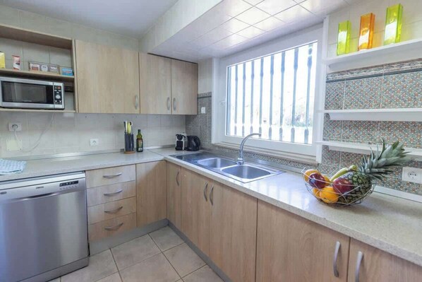 Una cocina con armarios de madera, lavavajillas de acero inoxidable, microondas, fregadero y ventana con persianas.