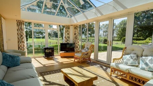 A spacious living room with a large sofa, a wooden coffee table, and two wicker chairs. A television is placed on a wooden stand, and the room has large glass doors leading to a garden. - The Cotswold Manor Vineyard, Exclusive Hot Tub, Games Barn, 70 acres of Parkland (Bampton)