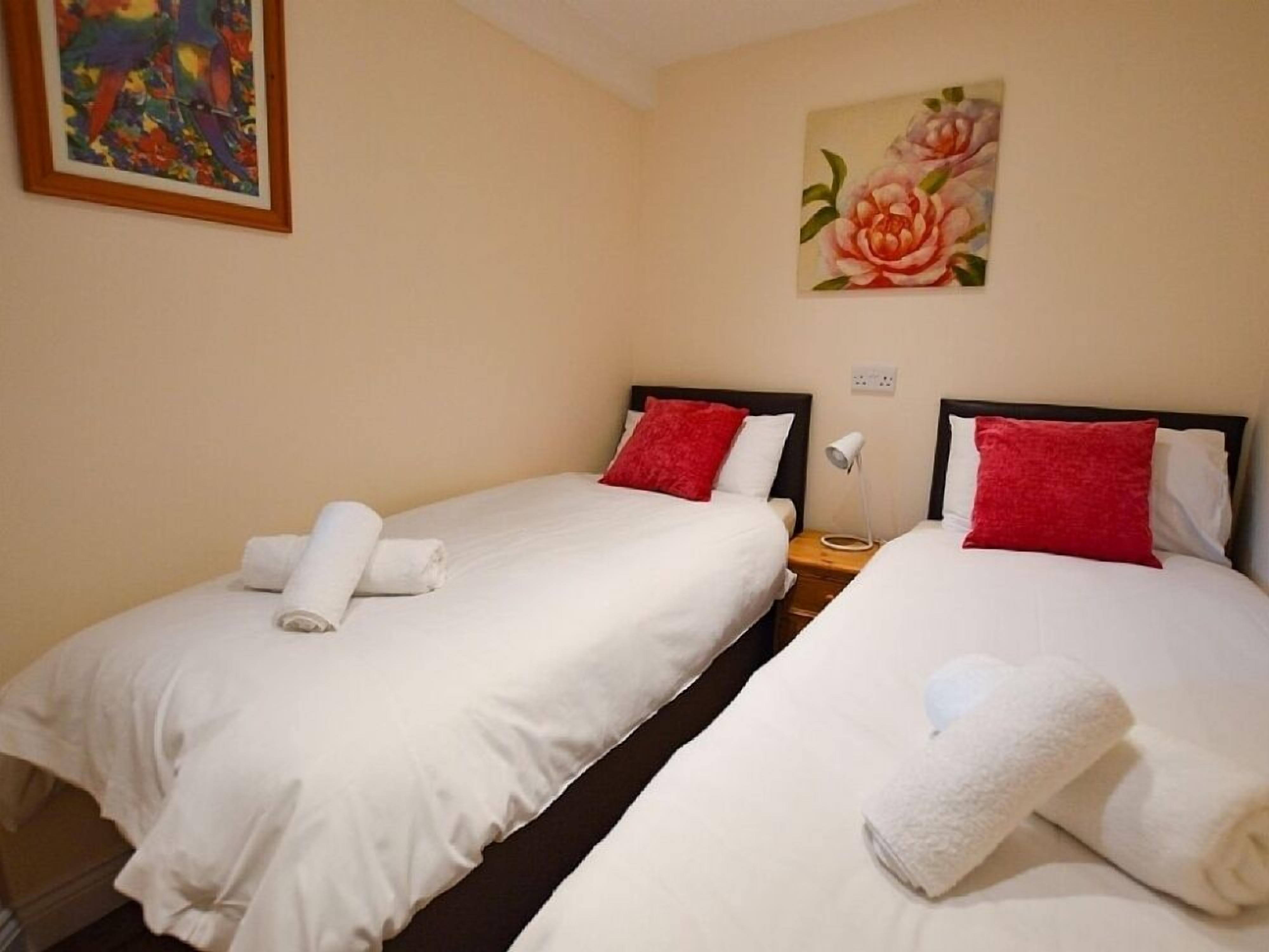 Dos camas individuales con sábanas blancas y almohadas rojas, un cuadro de flores en la pared y una mesita de noche con una lámpara entre las camas.
