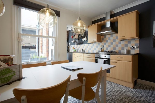 A kitchen with a white table, wooden chairs, hanging pendant lights, and a tiled backsplash.