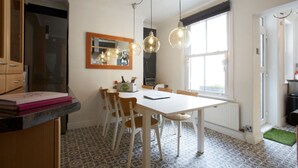 Een keuken met een witte tafel, houten stoelen, hanglampen, een raam en vloertegels met een patroon.