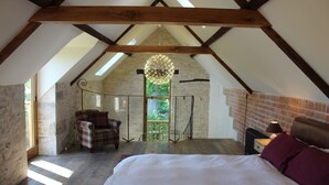 Un dormitorio en el ático con techo inclinado, un sillón de cuadros, cama con ropa de cama blanca y una pared de ladrillo.