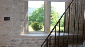 Una escalera con paredes de piedra y techo de vigas de madera, que conduce a un ventanal con vistas a la vegetación.