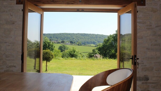 Una habitación con una amplia mesa de madera y una sola silla, una puerta corrediza de vidrio que da a una vista panorámica al aire libre de un paisaje verde y colinas.