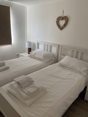 A hotel room with two single beds, white bedding, a heart-shaped wall decoration, a bedside lamp, and a window with blinds.
