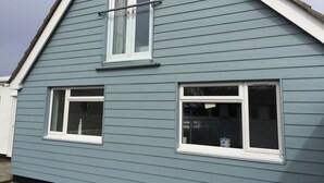 Une maison à deux étages, avec un toit à pignon, des boiseries blanches et deux fenêtres à cadres blancs.