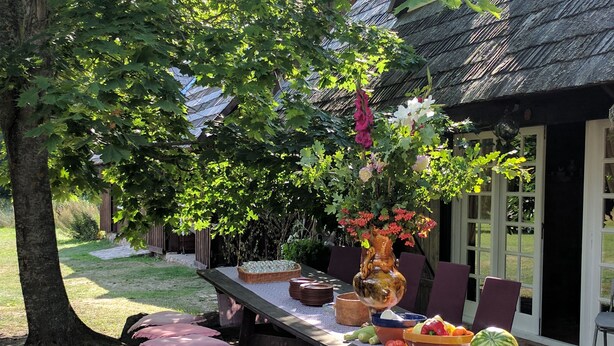 Une table en bois, installée à l’extérieur, est agrémentée d’un vase de fleurs, d’une pastèque, ainsi que de divers fruits et légumes.