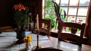 Une table en bois recouverte d’une nappe blanche, une bougie, un vase en céramique blanche et des plantes en pot.
