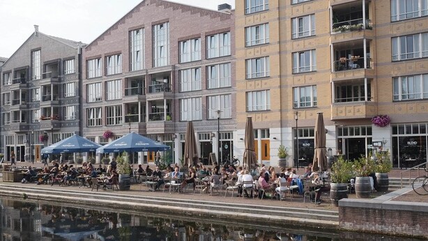 A riverside café with outdoor seating, a canal in the foreground, and a row of multi-story buildings with balconies. - Lovely spacious, luxurious apartment with canal view (Amsterdam)