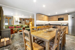 A modern kitchen with wooden cabinets, a stainless steel refrigerator, and a dining area with a wooden table and chairs. - Chalet Grand Cru Central 5 Bed with Jacuzzi (Chamonix-Mont-Blanc)