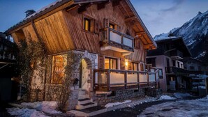 A wooden chalet with a balcony, stone foundation, and a mountain backdrop. - Chalet Grand Cru Central 5 Bed with Jacuzzi (Chamonix-Mont-Blanc)