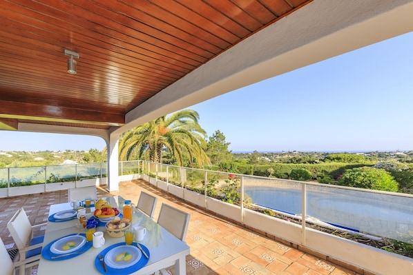 A covered outdoor dining area with a table set for breakfast, overlooking a swimming pool and lush greenery.