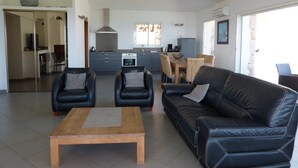 Una sala de estar moderna con un sofá de cuero negro, dos sillones y una mesa de centro de madera. Se ve una cocina con estufa, horno y mesa de comedor al fondo.