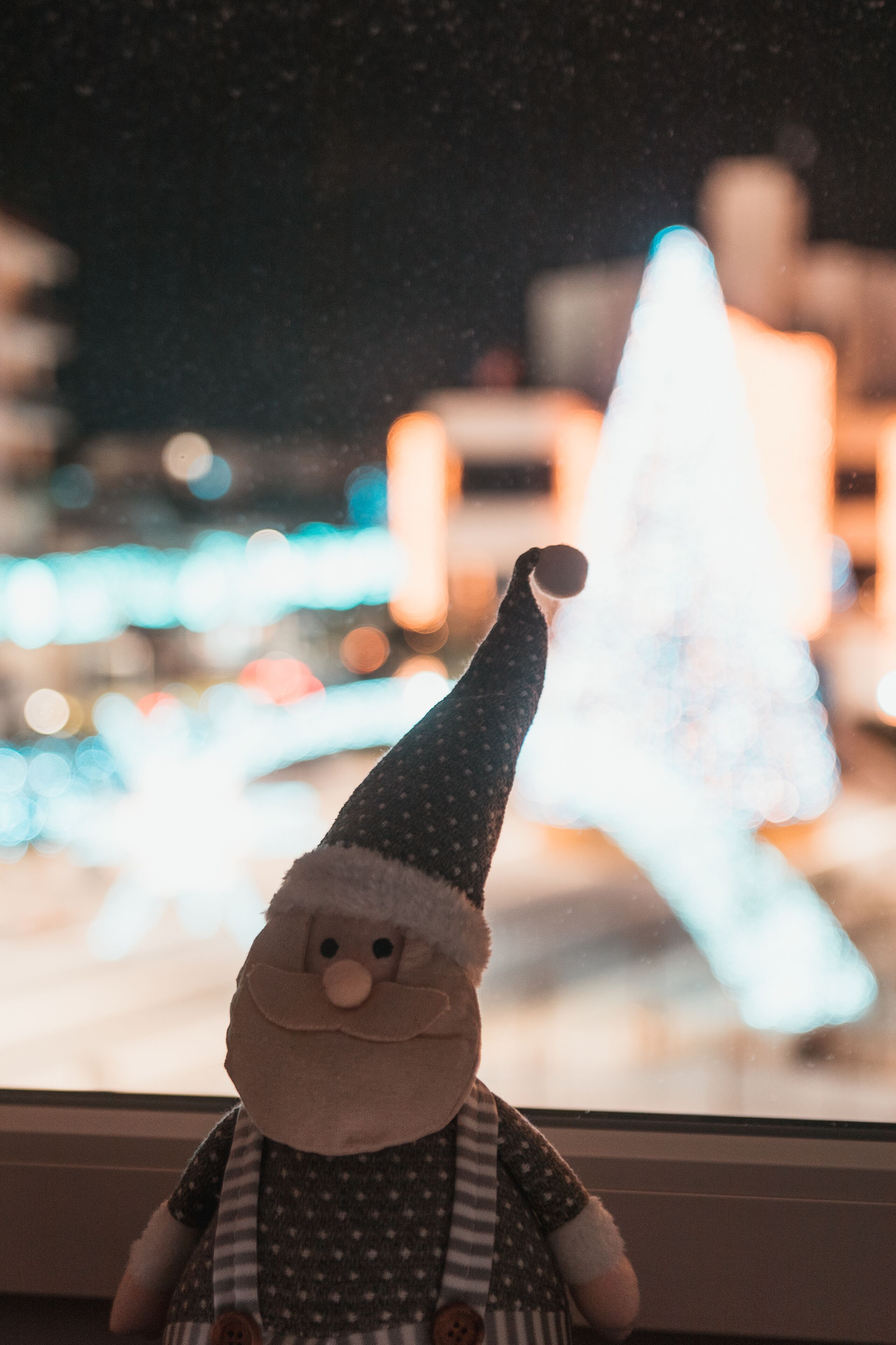 Un peluche de Papá Noel frente a una ventana, con un árbol de Navidad y luces desenfocadas al fondo.
