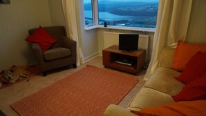 A living room with a sofa, a chair, a television on a stand, a patterned rug, and a dog lying on the floor. - Brynsior, Chalet on the hill in Goodwick with stunning sea views (Fishguard)
