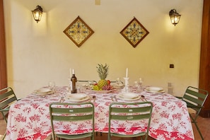 A dining table set for six with a floral tablecloth, two wall-mounted lanterns, and a decorative tile above the table.
