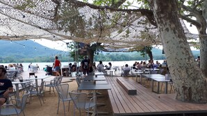 Ein Essbereich im Freien mit Tischen und Stühlen, eine Holzterrasse und Blick auf den See.