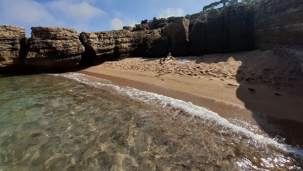 Ein Strand mit klarem Wasser, felsigen Klippen und einer Person, die im Sand liegt.