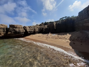 Ein Strand mit klarem Wasser, felsigen Klippen und einer Person, die im Sand liegt.