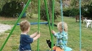 Dos niños jugando en un columpio en un área verde al aire libre, con árboles y una silla de fondo.