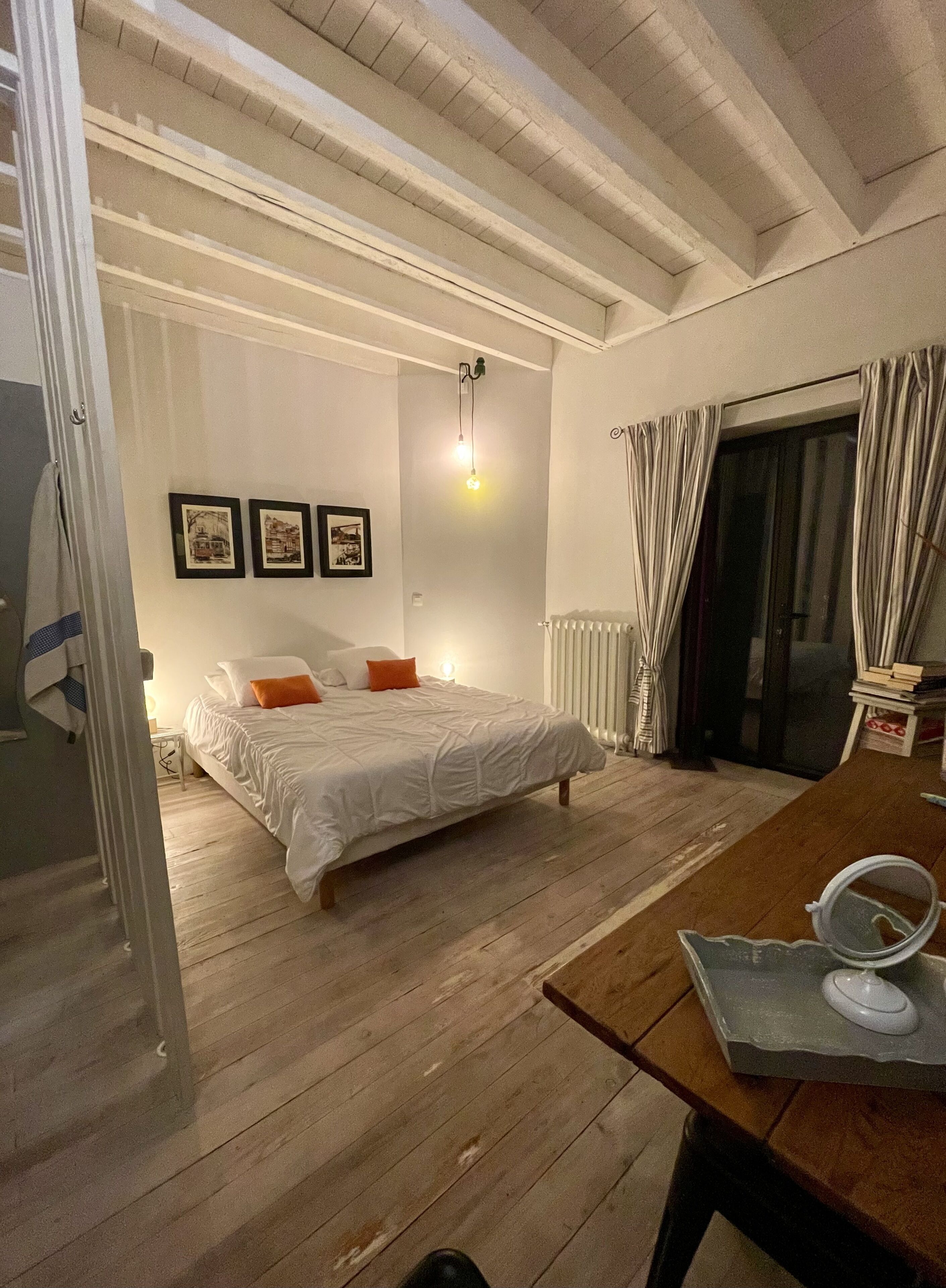 A bedroom with a bed, a wooden floor, a table with a mirror, and a window with curtains.