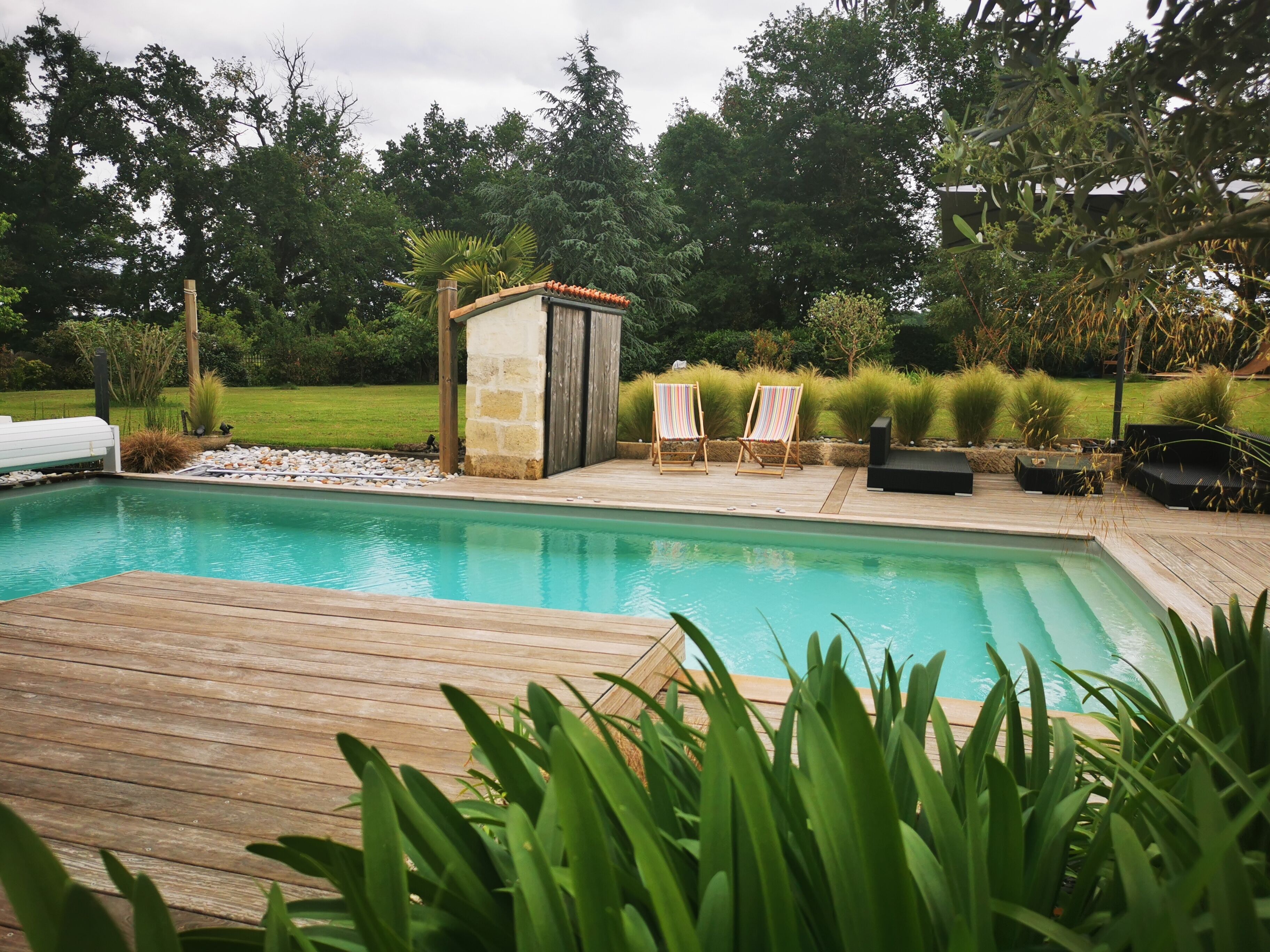 Una piscina con cubierta de madera, rodeada de exuberante vegetación y dos sillas de descanso.