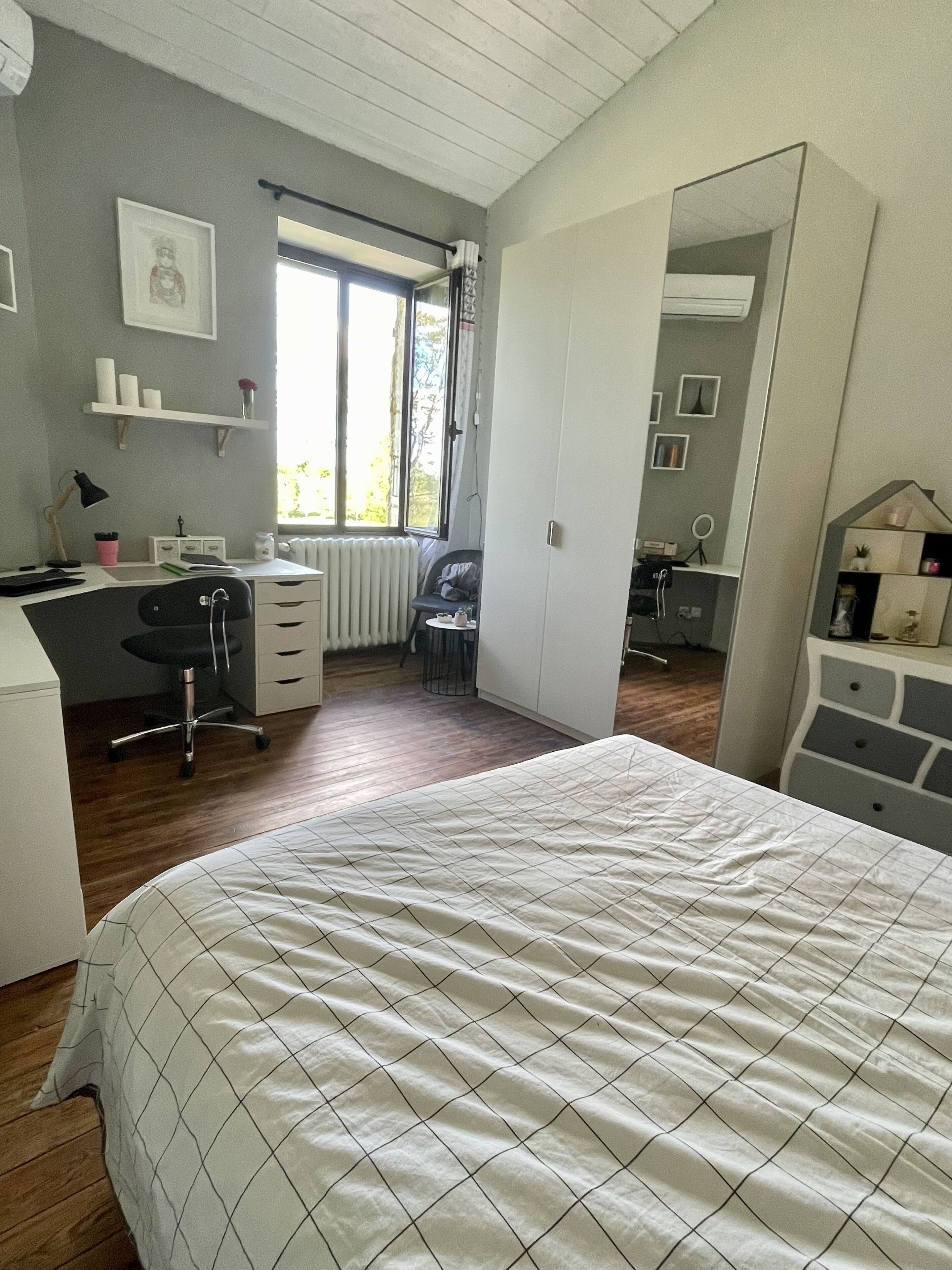 Un dormitorio con cama, escritorio, silla, radiador, ventana y espejo.