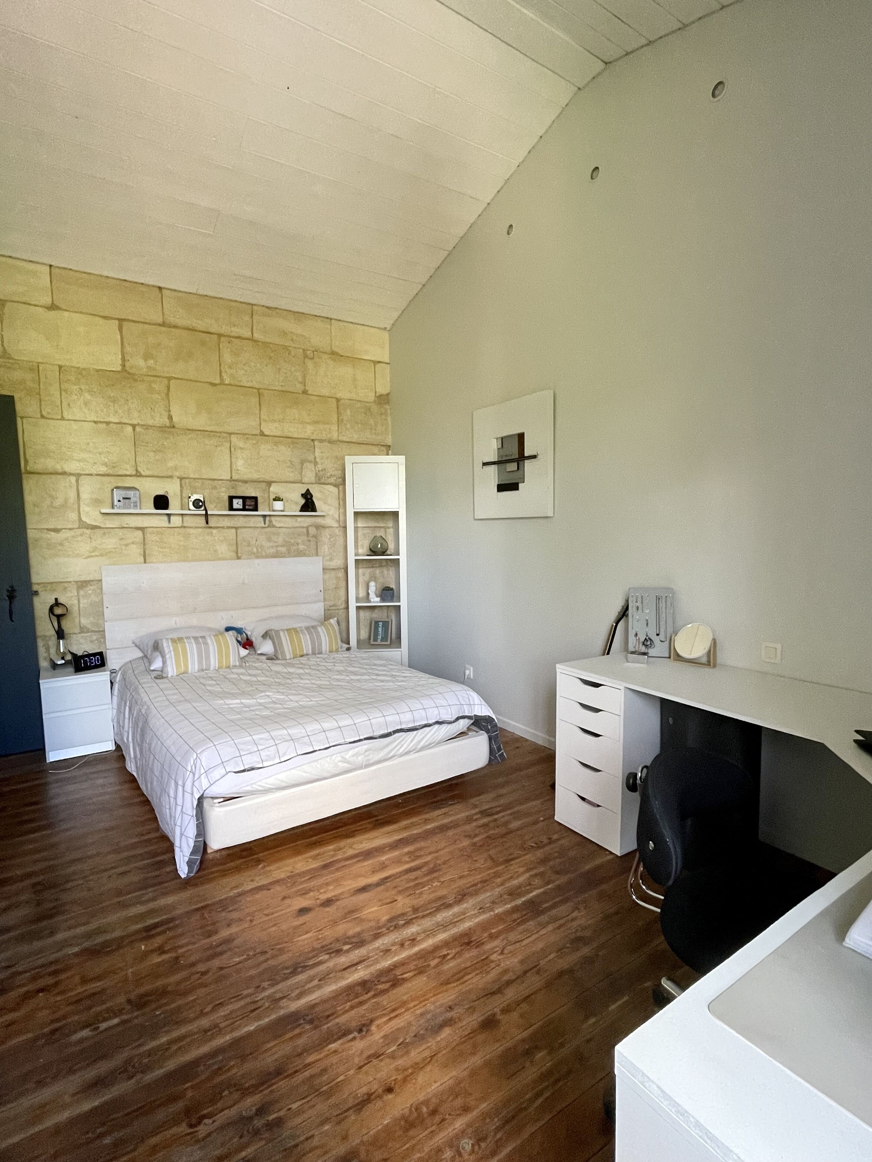 Een slaapkamer met een bed, nachtkastje, dressoir, spiegel en plank met diverse spullen.
