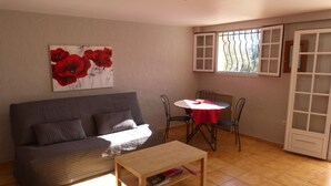 Een woonkamer met een grijze bank, een houten salontafel en een eettafel met stoelen. Er is een raam met metalen tralies en een deur met ruitpanelen.