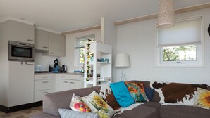 A modern living room with a sofa, a coffee table, a kitchen area with appliances, and a window with blinds. - Chalet Zonnebloem for 6 people, lots of privacy in a quiet park (Serooskerke (W))