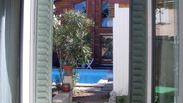 Une vue à travers une fenêtre dont les volets sont ouverts, dévoilant une piscine, une maison en bois et un petit jardin.