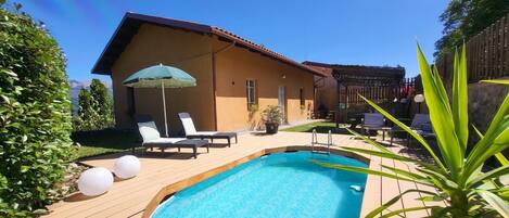 A swimming pool with lounge chairs and umbrellas, surrounded by a wooden deck and a building with a sloped roof.